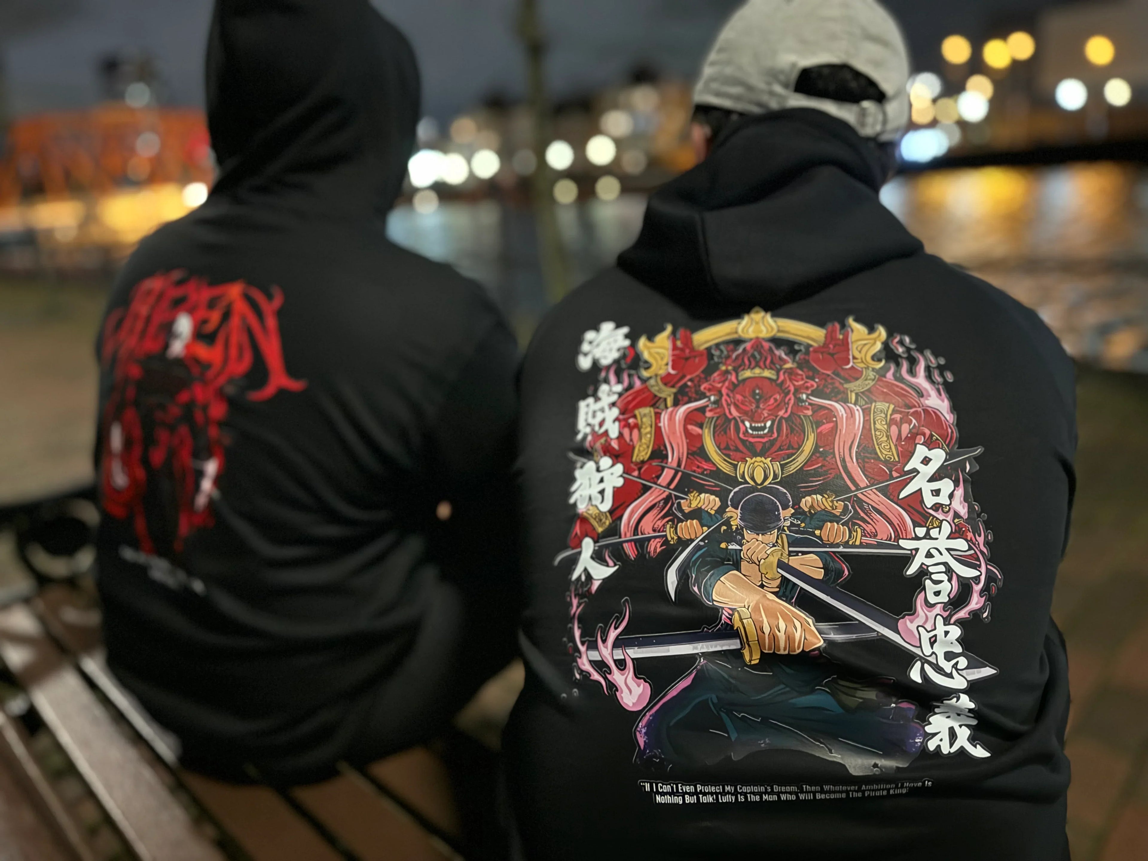 Two men sitting by the water at night wearing black Kunai anime hoodies, showing large detailed back graphics with Japanese Anime artwork