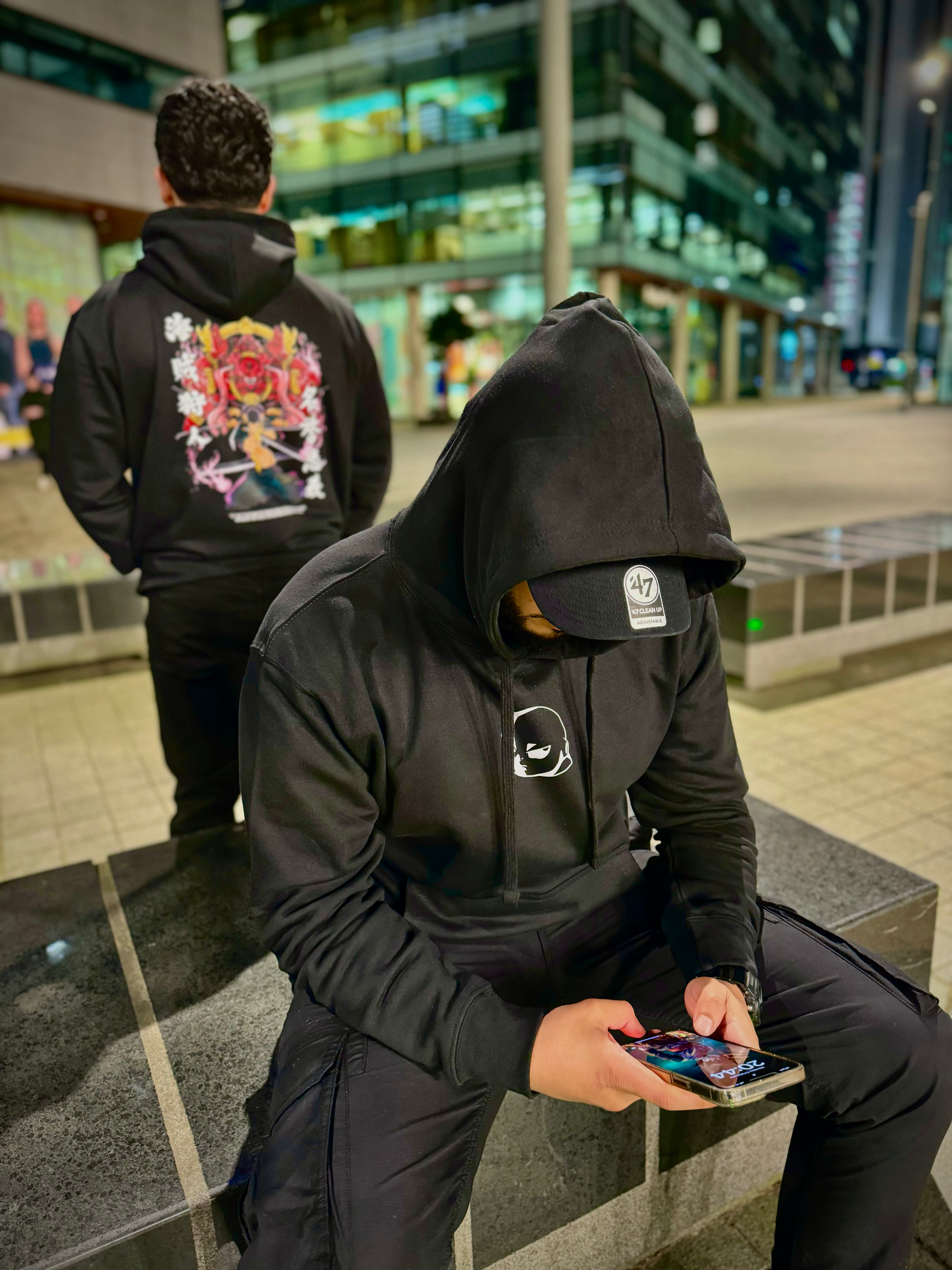 Two individuals wearing black hoodies with colorful designs in an urban setting.